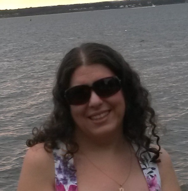 Headshot of Mikaila standing in front of a body of water, wearing sunglasses and smiling, wearing white with flowers.